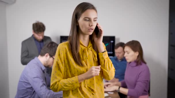 Jonge brunette vrouw is praten door mobiele telefoon staan in de vergaderzaal van kantoor, werknemers zijn op de achtergrond — Stockvideo