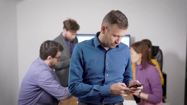 Homem está digitando sms no smartphone, de pé na sala de reuniões do escritório, pessoas de negócios estão se comunicando — Vídeo de Stock