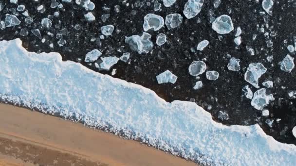 Top aerial view from above. A beautiful designer view of the banks of the freezing river. The sandy beach rests on the edge of the ice, then individual ice floes float in cold water. — Stock Video