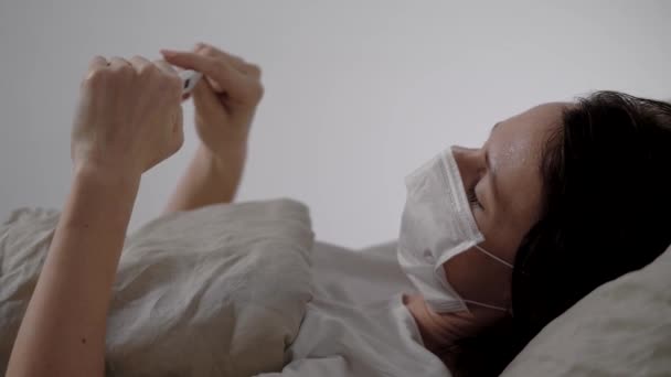 Una chica enferma está en la cama. El paciente mide su temperatura con un término electrónico momentum. Epidemia — Vídeo de stock