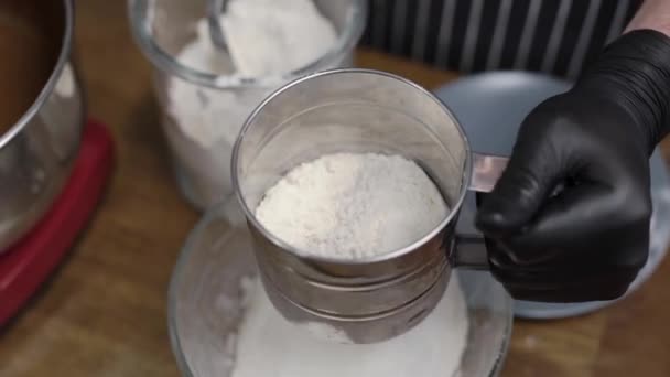 El pastelero tamiza a través de la harina. En el marco de las manos en guantes negros con un sito. Cocinar — Vídeos de Stock