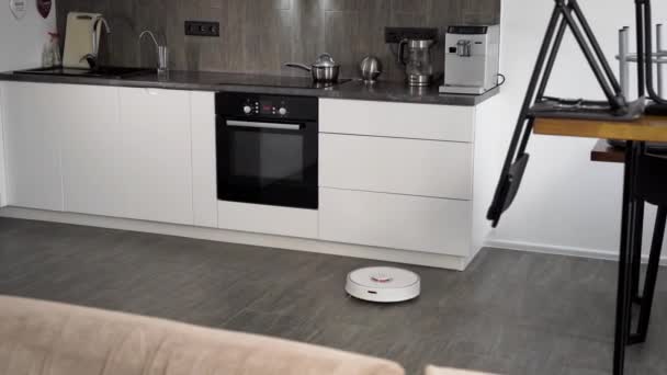 Em uma cozinha moderna elegante é a limpeza. O aspirador de pó robô automático se move ao longo de sua trajetória. Elementos de uma casa inteligente tornam a vida mais fácil para as pessoas — Vídeo de Stock