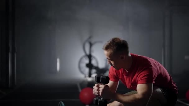 Sportsman is squatting with dumbbells in hands in dark gymnastic hall, training legs and hands, keeping fit — Stock Video