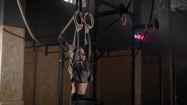 Joven mujer está colgando de anillos deportivos en sala de gimnasia y levantamiento de piernas, tensando los músculos del abdomen — Vídeos de Stock