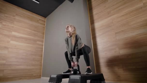 Fille athlétique exécute squats dans salle de fitness, tenant kettlebell dans les mains, debout sur les plates-formes — Video