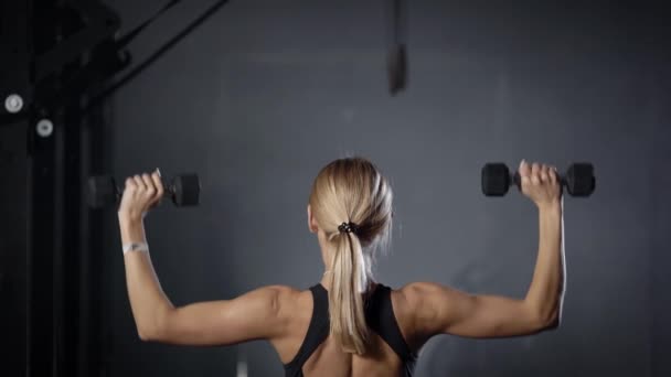 Спортивная молодая блондинка поднимает гантели руками в спортзале, стоя спиной к камере — стоковое видео