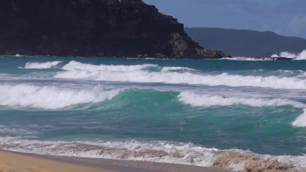 Gyönyörű óceáni hullámok hemperegnek a homokos parton. A sziklák és hegyek hátterében. — Stock videók