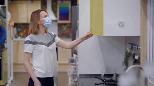 Una hermosa joven con una máscara protectora. La chica elige la cocina en su nuevo hogar . — Vídeos de Stock