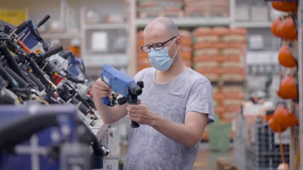 Un uomo con una maschera protettiva e occhiali sceglie un trapano. Un operaio di un negozio di utensili elettrici vuole comprare un puncher . — Video Stock