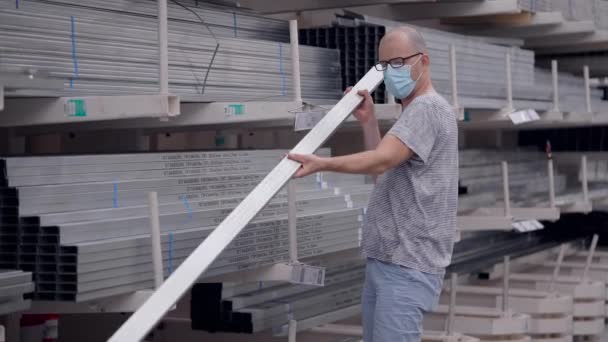 Un uomo con una maschera protettiva ha visitato un negozio di costruzioni. Il rabochy sceglie un profilo metallico per il telaio . — Video Stock