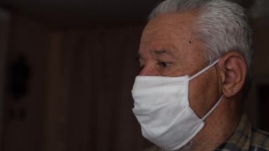 Evde cerrahi maske takan kel ve olgun bir adam video konferans görüşmesi için akıllı telefon kullanıyor. Koronavirüs SARS-CoV-2 salgını ve COVID-19 nedeniyle sosyal mesafeler.