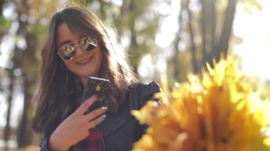 Genç bir kadın telefon kamerasında sonbahar yapraklarının fotoğrafını çekiyor. Parlak sarı yapraklar..