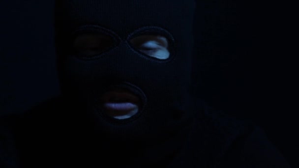 Мужчина в балаклаве в темной комнате смотрит в камеру — стоковое видео