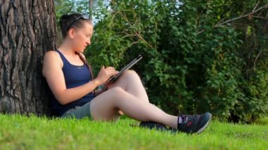 Parktaki ağacın altında yeşil bir çimende oturan genç kadın tablet kullanıyor.