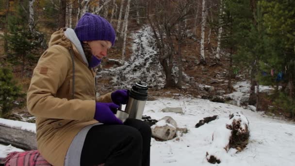 Young woman pours tea from a thermos near a stream in the winter forest — Stock Video