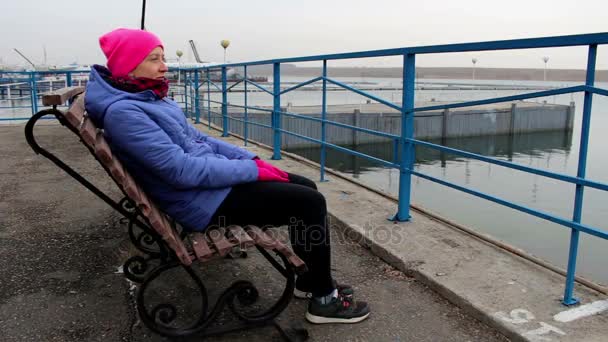 Chica sentada en un banco en el muelle en un frío día nublado y sonriendo — Vídeo de stock