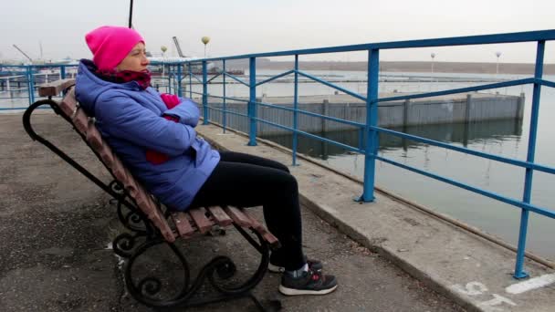 Tjejen sitter på en bänk på bryggan en kall mulen dag ser i fjärran — Stockvideo