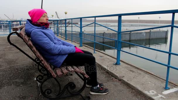 Menina sentada em um banco no cais em um dia nublado frio olha para a distância — Vídeo de Stock