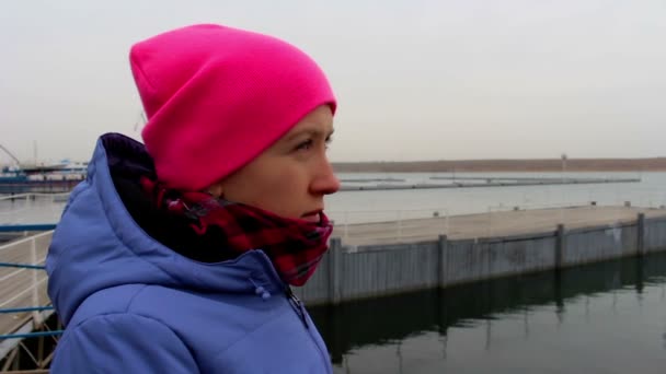 Retrato de una niña con un sombrero rosa en el muelle en un día frío y nublado en otoño — Vídeo de stock