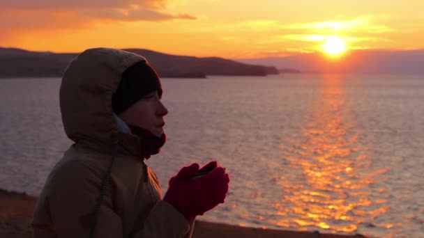 La chica se sienta en la orilla del mar y bebe un té caliente. Hermoso atardecer dorado — Vídeo de stock