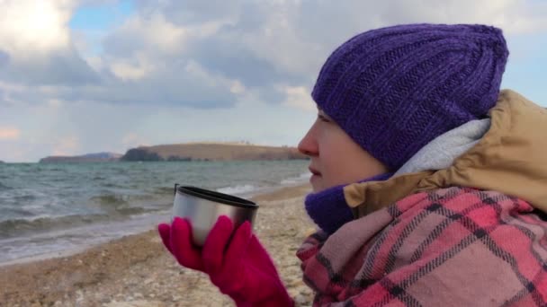 La chica se sienta en la orilla del mar y bebe un té caliente. Olas, hermosos paisajes — Vídeos de Stock