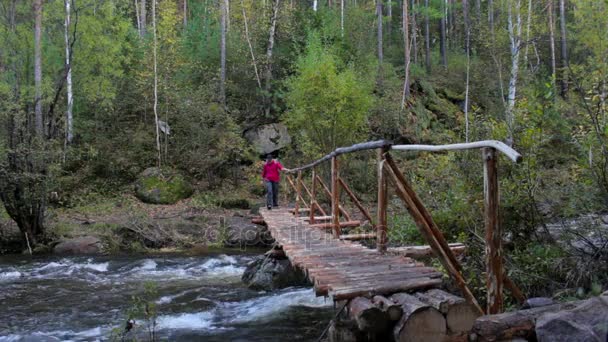 Turystyczna dziewczynka z plecakiem idzie na drewniany most na górskiej rzece — Wideo stockowe
