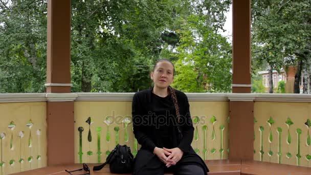 Porträtt av en leende flicka med fläta klädd i svart sitter i lusthuset — Stockvideo