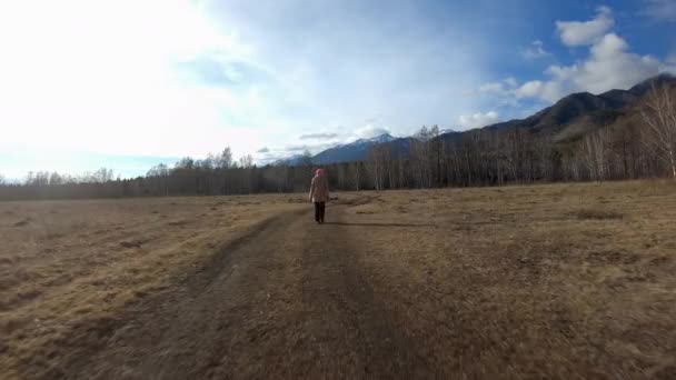 Κορίτσι περπατά κατά μήκος ενός επαρχιακού δρόμου σε ένα χωράφι στους πρόποδες των βουνών το φθινόπωρο — Αρχείο Βίντεο