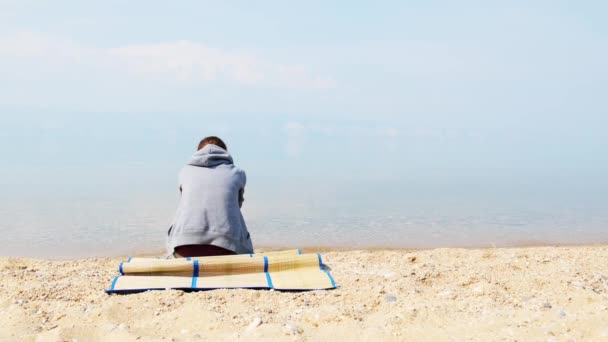 Una chica triste y solitaria está sentada en la playa con una sudadera gris. Vista trasera — Vídeo de stock