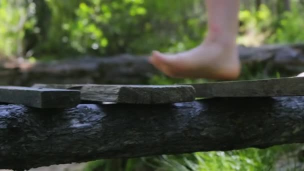 Ένα κορίτσι με γυμνά πόδια περπατάει σε μια ξύλινη γέφυρα σε ένα καταπράσινο δάσος. Κοντινό πλάνο ποδιών — Αρχείο Βίντεο