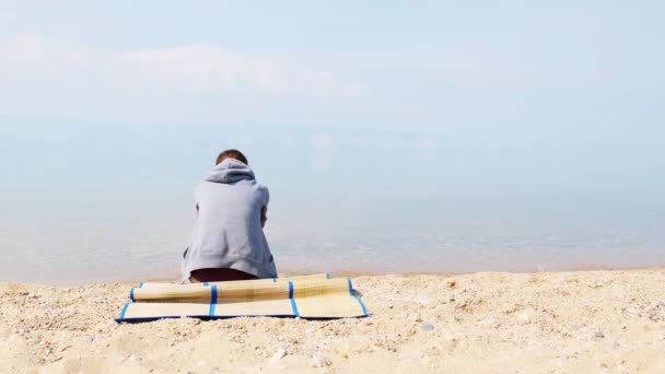 Ένα μοναχικό κορίτσι κάθεται στην παραλία φορώντας γκρι κουκούλα. Πίσω όψη — Αρχείο Βίντεο