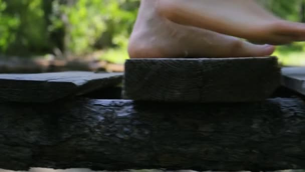 Jambes rapprochées. Une fille aux pieds nus marche sur un pont en bois dans une forêt verte — Video