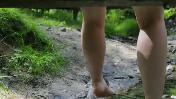 Fille s'assoit sur un pont en bois sur un ruisseau et pend ses pieds nus dans l'eau — Video