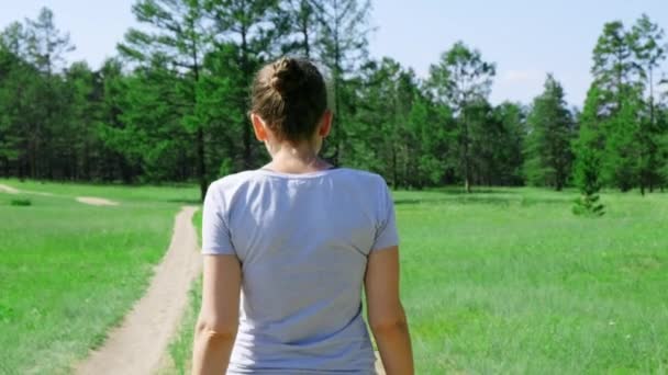 Uma menina caminha ao longo de uma estrada rural em um prado verde cercado por floresta — Vídeo de Stock