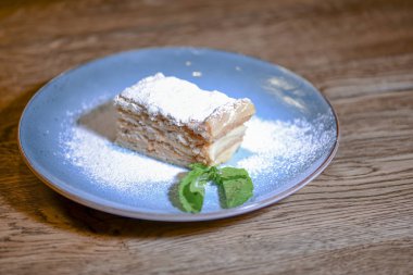 Kremalı pasta Napolyon, nane şekerli vanilya dilimi ve ahşap masada mavi tabakta servis edilen şeker tozu..