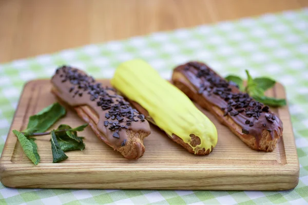 Traditional french dessert eclair pastry, three pieces laying on wooden board on a table covered with plaid fabric.