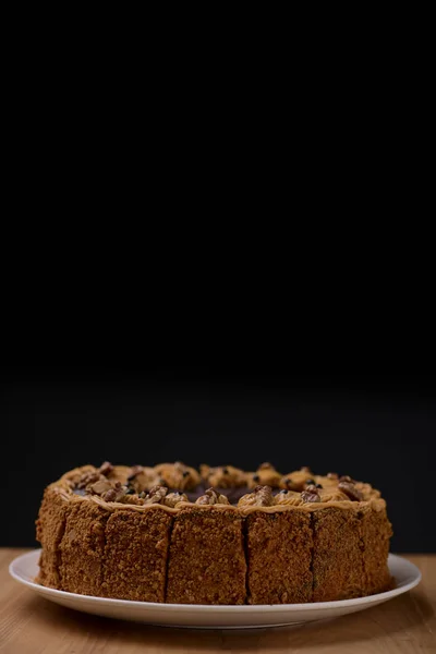 Delicious nut cake glazed with dark chocolate icing served on a white plate on light wooden table background.