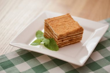Bir dilim katmanlı vanilyalı bal keki beyaz bir tabakta, tahta masada nane yaprağı ve ekose masa örtüsü.