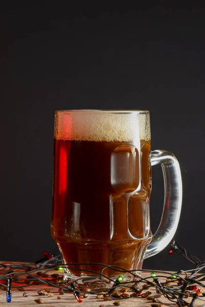 Vaso completo de oso o cerveza con luces navideñas sobre una mesa de madera clara sobre fondo negro. Cerveza fuerte, cerveza de trigo . — Foto de Stock