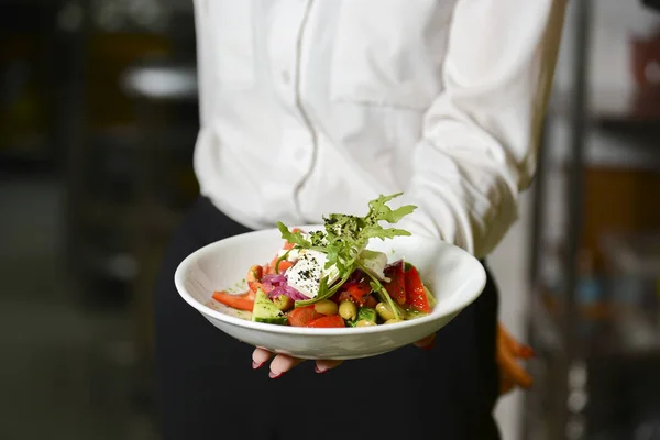 Waiter holds a plate with tasty dish, traditional Italian Greek salad. Waiter wearing white uniform in a restaurant.