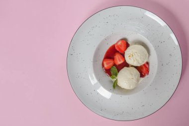 Çilekli vanilyalı çilekli dondurma ve pastel pembe arka planda beyaz bir tabakta meyve şurubu. Üst görünüm.