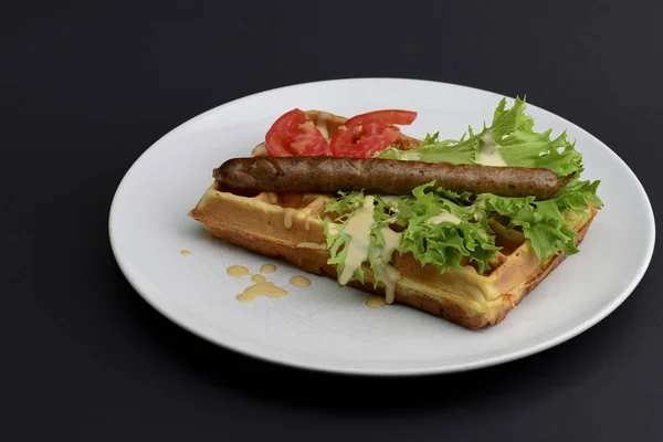 Saucisse classique sur gaufres petit déjeuner avec tomates, salade et sauce servi sur une assiette blanche sur fond noir . — Photo