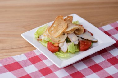 Geleneksel klasik Sezar salatası, beyaz bir tabakta, beyaz tahta bir masanın üzerinde, ekose masa örtüsüyle servis ediliyor..