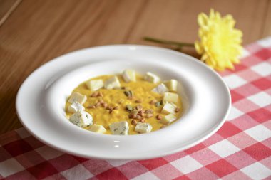 Kabak çekirdeği ve peynirle kaplı kremalı balkabağı çorbası ahşap bir masada, ekose masa örtüsüyle..