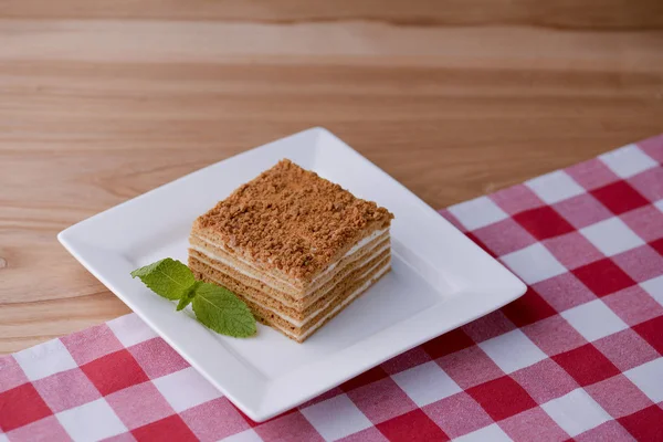 Una rebanada de pastel de miel de vainilla en capas en un plato blanco con una hoja de menta en la mesa de madera con un paño de tabla a cuadros —  Fotos de Stock