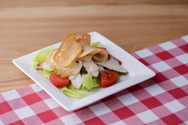 Традиционный классический салат Цезарь с курицей подается в белой тарелке над светлым деревянным столом с клетчатой скатертью . — стоковое фото