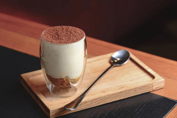 Traditional Italian dessert tiramisu in a glass served on a wooden board with a spoon in a restaurant. Italian cuisine