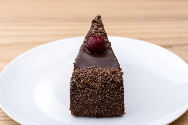 Kawałek ciasta czekoladowego z kremem waniliowym i polewą wiśniową na białym talerzu na jasnym drewnianym tle. — Zdjęcie stockowe