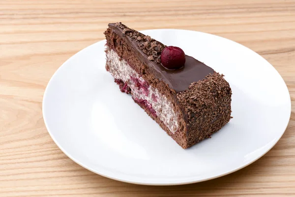 Um pedaço de bolo de chocolate com creme de baunilha e cobertura de cereja servido em placa branca sobre fundo de madeira leve . — Fotografia de Stock