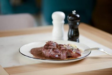 Lokantada beyaz tabakta ızgara biftek ve hafif ahşap masada çatal bıçak. Restoran yemek fotoğrafçılığı.